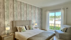A bedroom with a large bed, bedside tables, a chair, and a view of a green landscape through the window.
