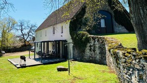 Une maison avec un mur en pierre, un espace extérieur aménagé et couvert, et un grand arbre au premier plan.