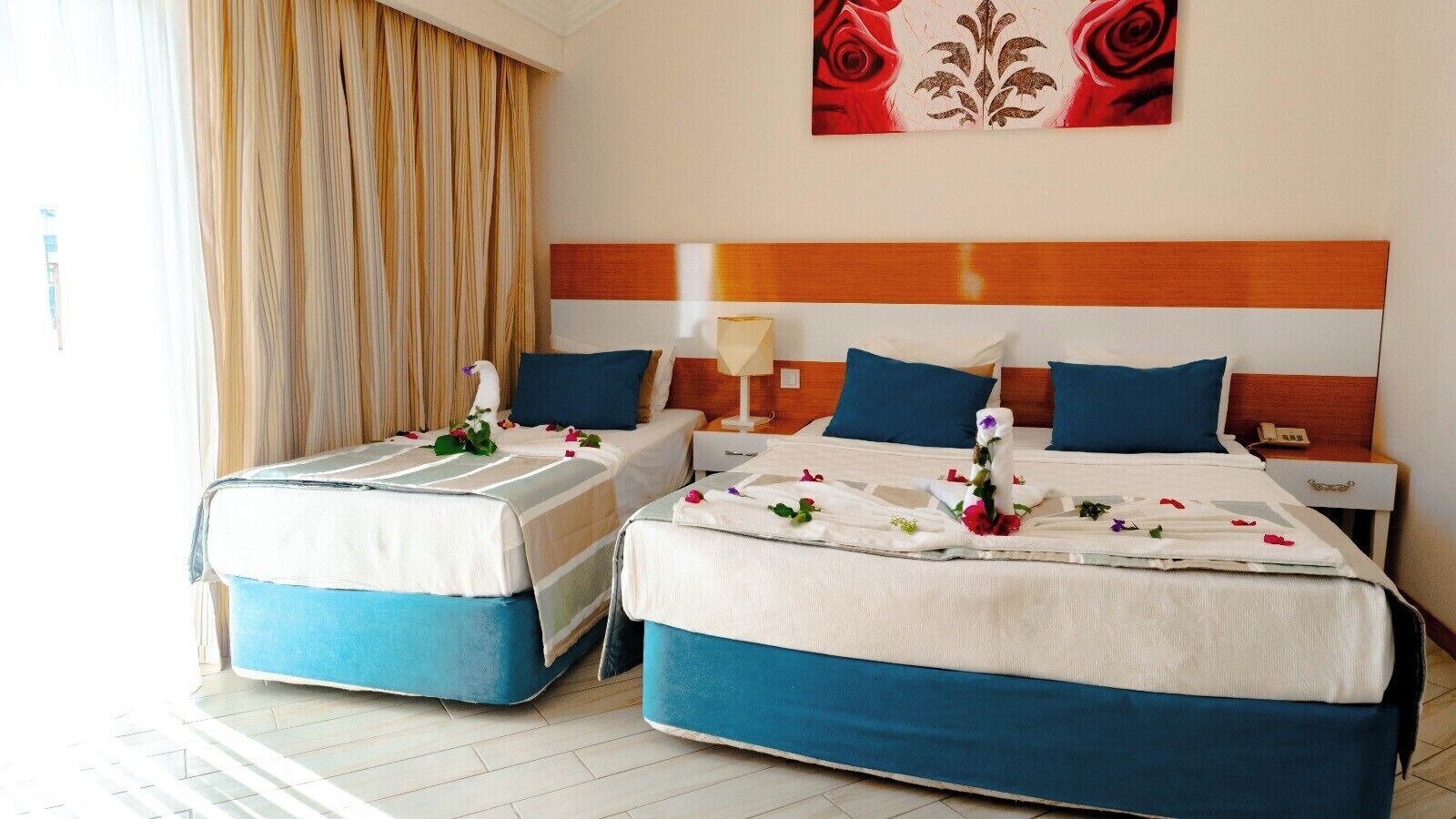 A hotel room with two beds, a nightstand, a lamp, and a painting of roses.