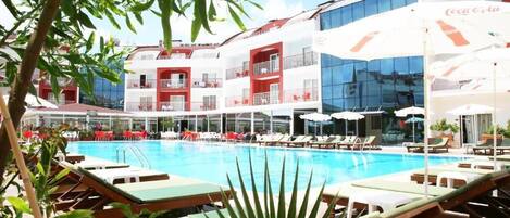 A hotel pool area with sun loungers, a Coca-Cola branded umbrella, and a glass building in the background.