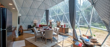 Interior de una cúpula geodésica con comedor, cocina y ventanas amplias que ofrecen vistas del exterior.