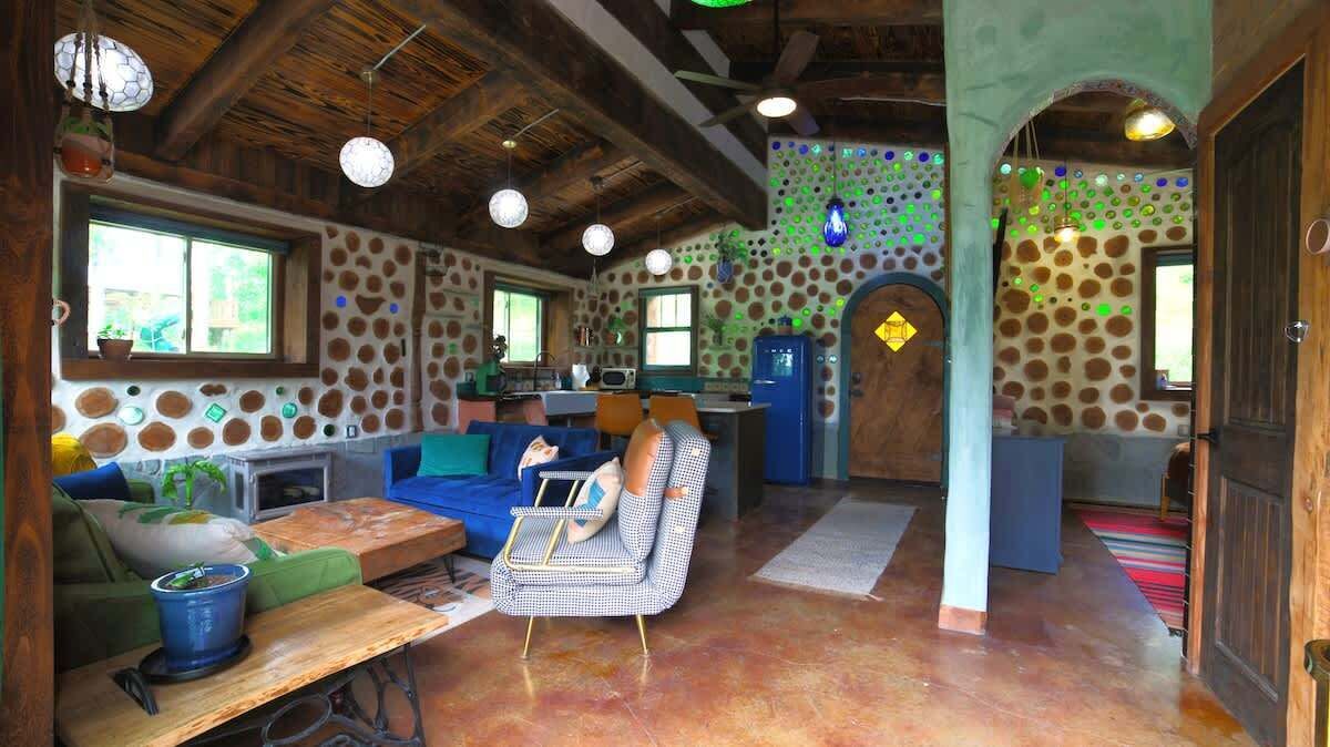A rustic interior with a stone wall, wooden beams, a blue sofa, a dining table, and a kitchen area.