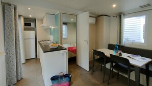 Una cocina compacta con estufa, lavamanos y microondas. Hay un comedor con una mesa y sillas, y una ventana pequeña con persianas.