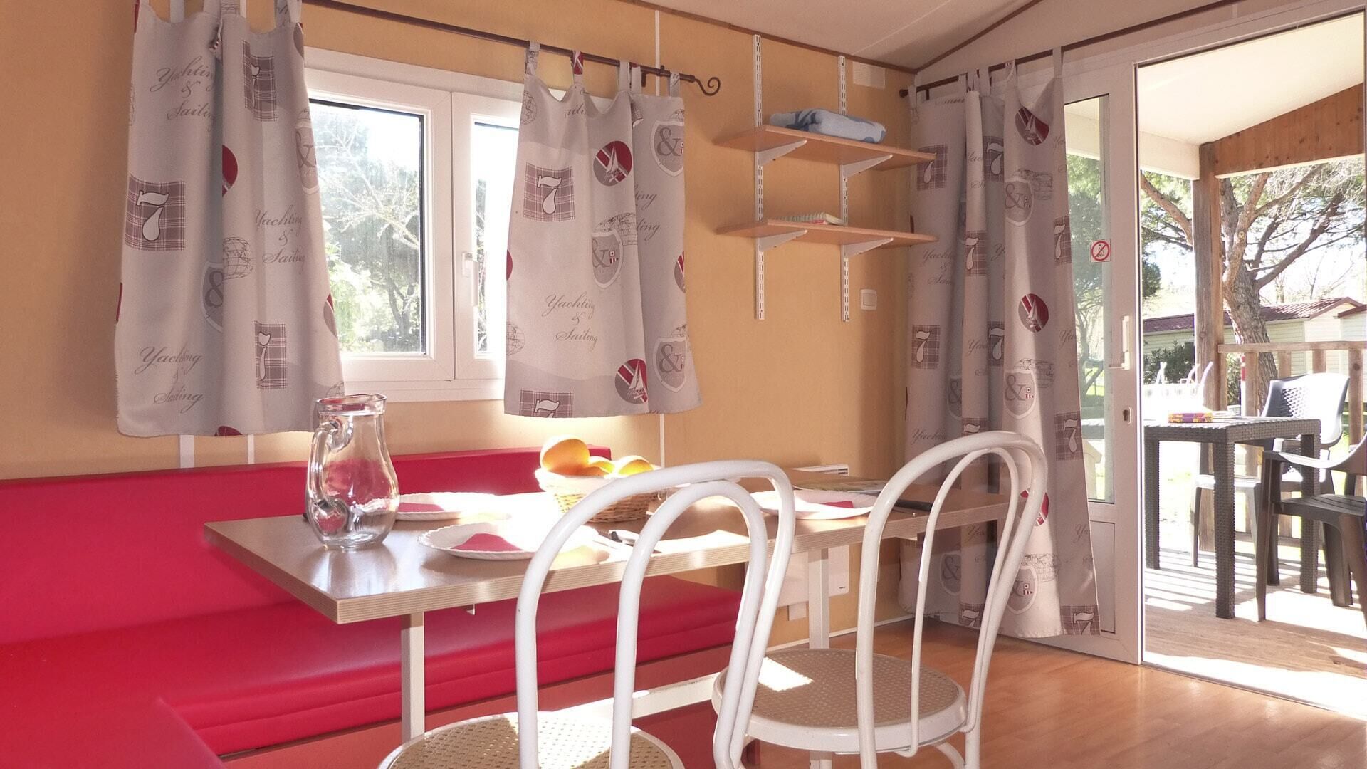 A dining area with a red bench, white chairs, and a table. There are curtains with a nautical theme, a window, and a view of the outside.