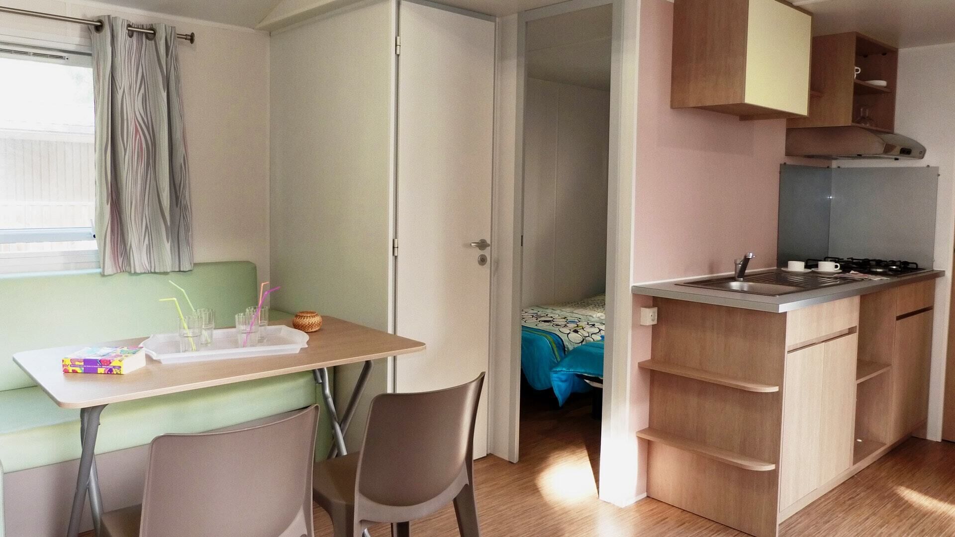 A compact kitchenette with a sink, stove, and wooden cabinets. There is a small dining area with a table and chairs, and a bed with a blue blanket in the background.