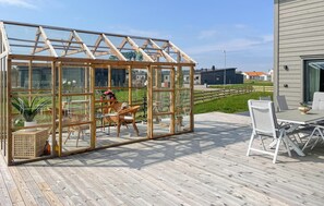 Una terrassa de fusta amb una estructura de vidre semblant a un hivernacle, mobles de vimet i vista cap a cases properes.