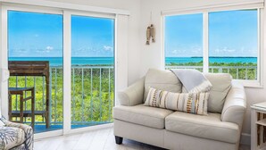 A room with a beige sofa, a wooden chair, and a view of the ocean through large windows. - Sunrise Oasis - Top Floor 2 BR 2 BA Condo  2411 (Tavernier)