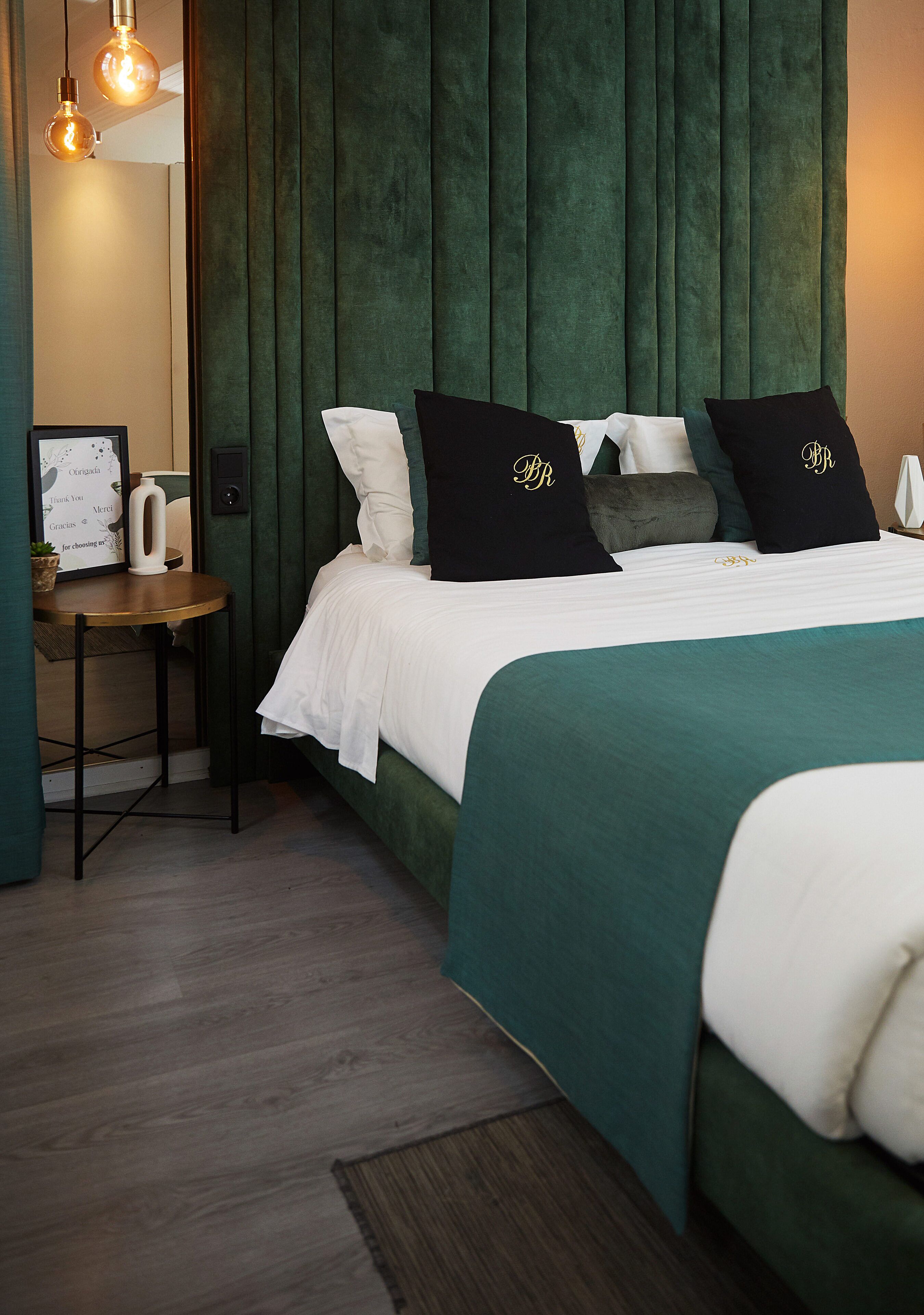A neatly arranged bedroom with a green upholstered headboard, white bedding, and black pillows with gold lettering.