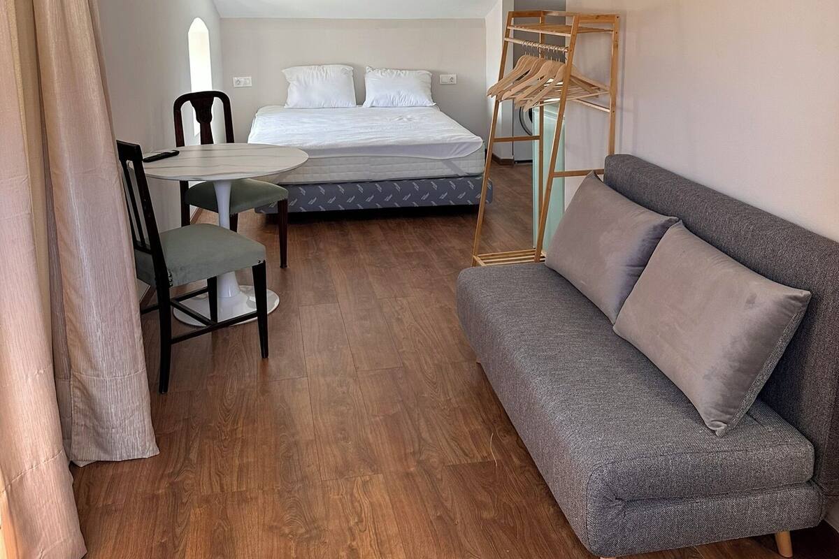 Une pièce moderne et bien éclairée, comprenant un lit, un canapé gris, une table à manger avec des chaises et un luminaire suspendu.