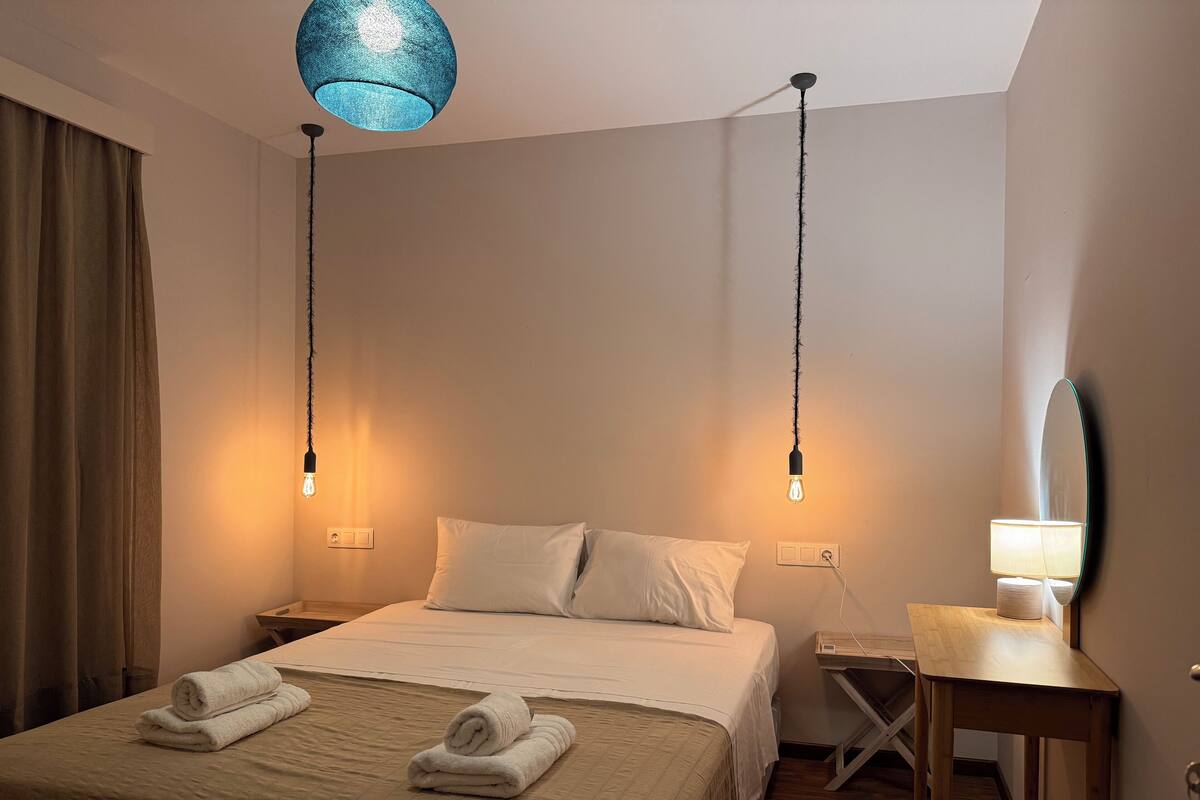 Une chambre d’hôtel avec un lit, deux lampes de chevet, un bureau avec une lampe, un miroir et un luminaire suspendu bleu.