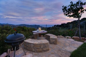 An outdoor patio with a stone fire pit, a grill, and a view of distant hills. - Holiday villa close to Triopetra beach, South Crete (Triopetra, Rethimno)