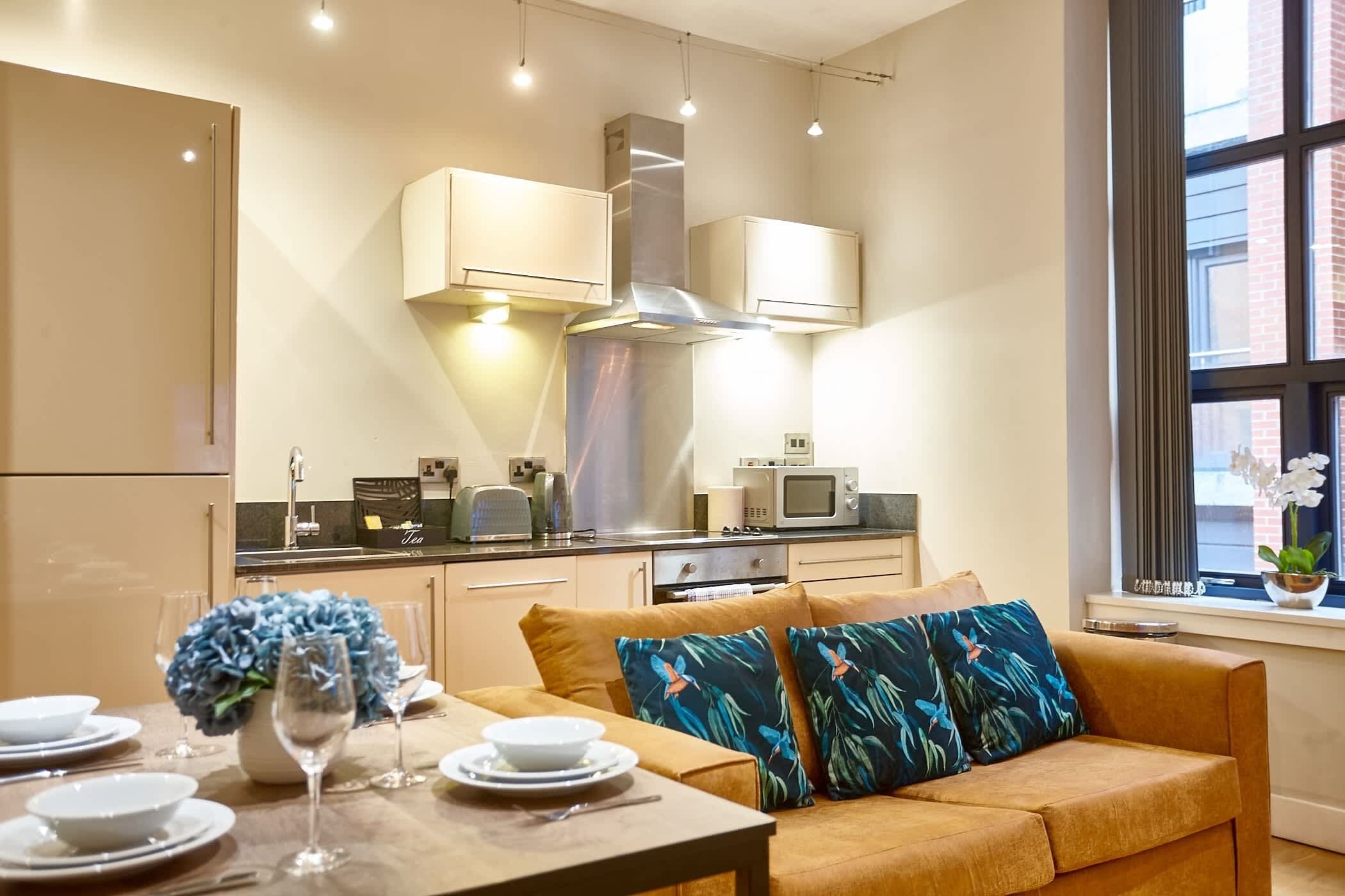 Een moderne keuken met een inbouwmagnetron, fornuis en oven. Er is een eethoek met een tafel voor twee personen, een bank met decoratieve kussens en een raam met uitzicht op de stad.