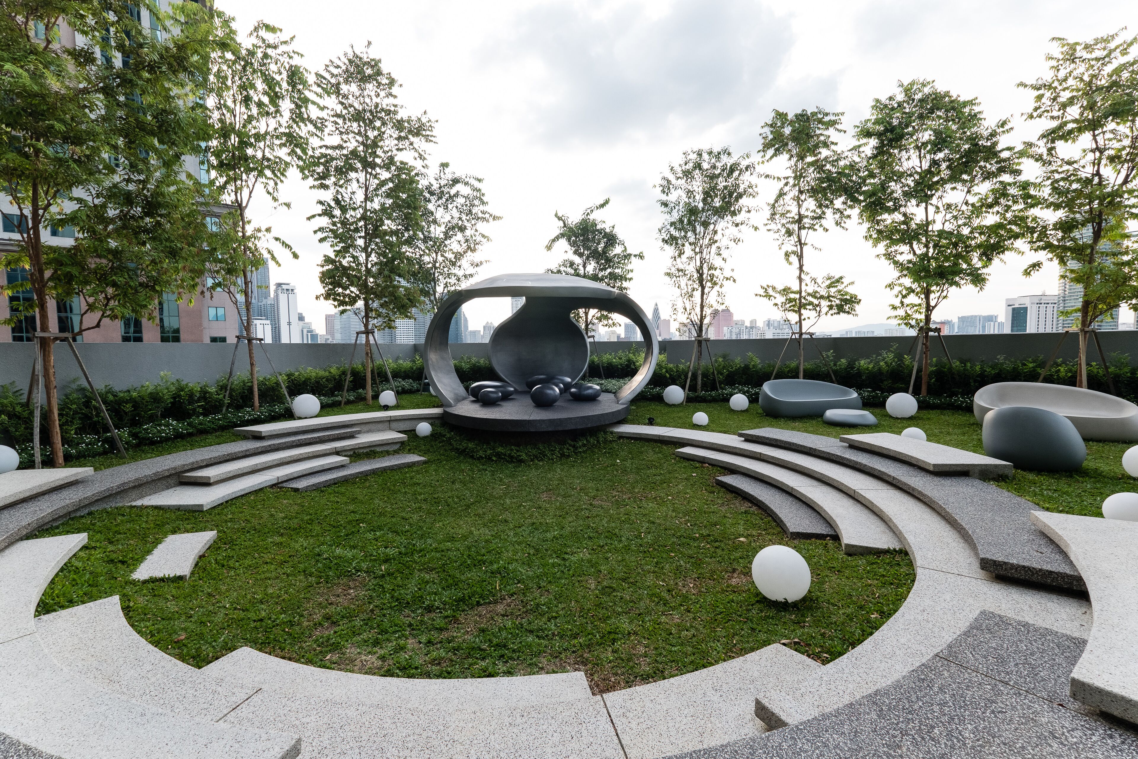 A modern outdoor space with a central circular seating area, surrounded by a landscaped garden with trees and a cityscape in the background.
