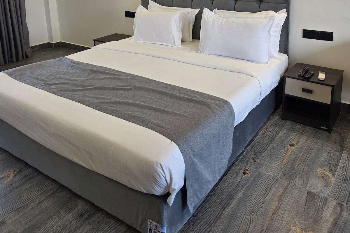 Una habitación de hotel moderna con una cama grande, ropa de cama gris y suelo de madera. Hay dos mesitas de noche con lámparas y un mueble auxiliar con teléfono y control remoto.