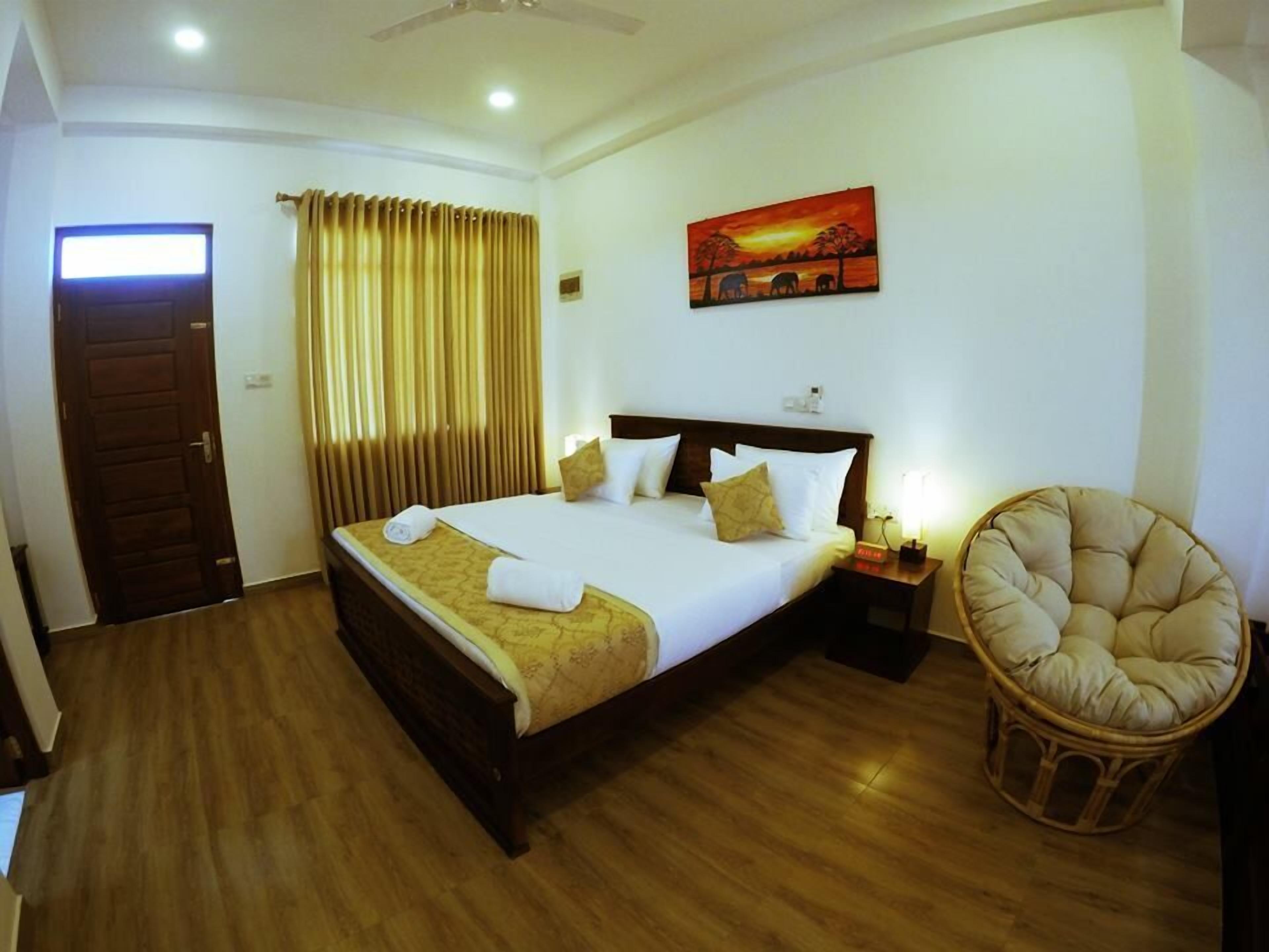 A hotel room with a wooden floor, a bed with white linens, a headboard, a nightstand with a lamp, a chair, a painting on the wall, and a door.