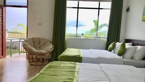 A hotel room with two beds, a wicker chair, and a balcony with a view of greenery.