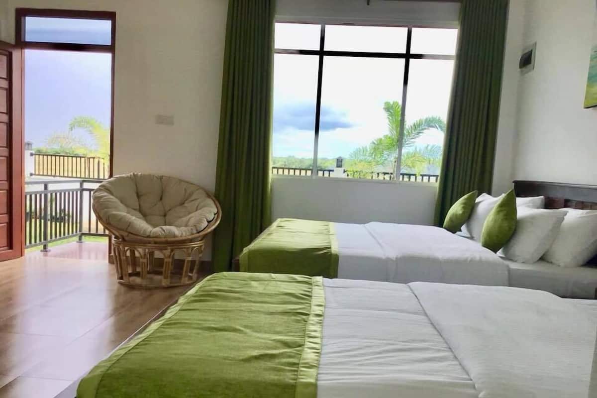 Habitación de hotel con dos camas, una silla de mimbre y un balcón con vistas al verdor.
