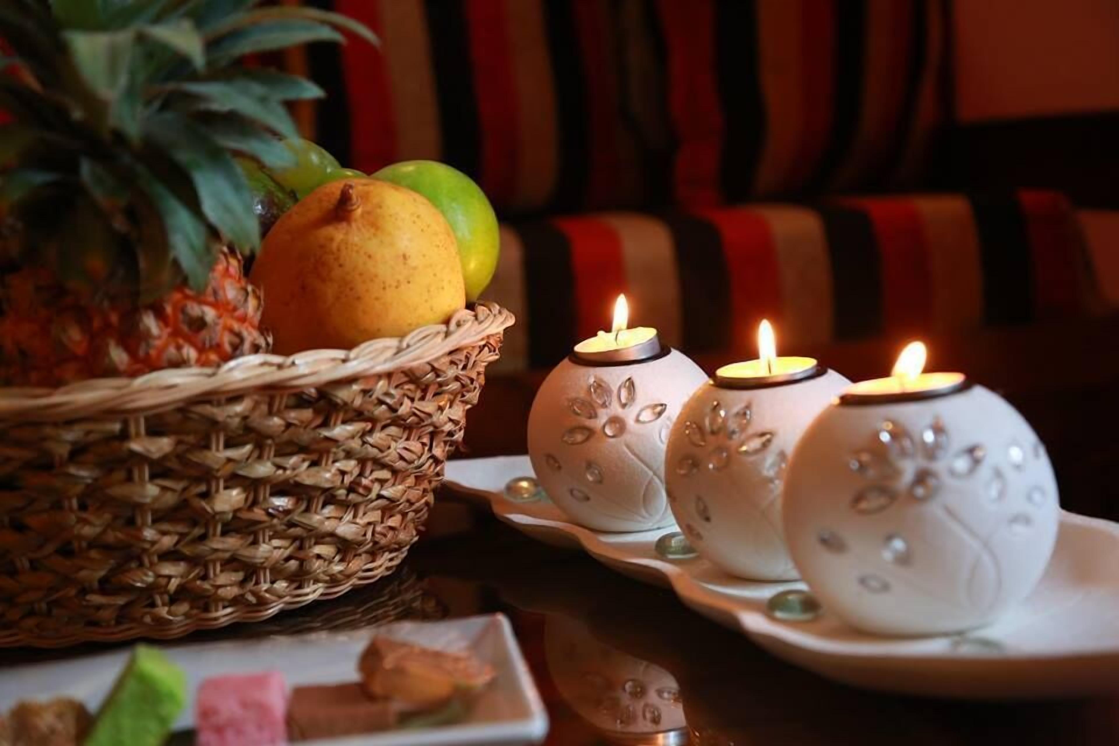 Una mesa con una cesta tejida llena de frutas, velas encendidas en jarrones decorativos y un plato con comida.