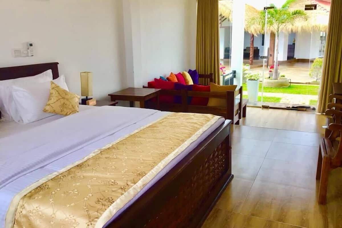 Habitación de hotel con una cama grande, una mesita pequeña, un sofá con cojines de colores y vistas a un espacio tropical con palmeras.