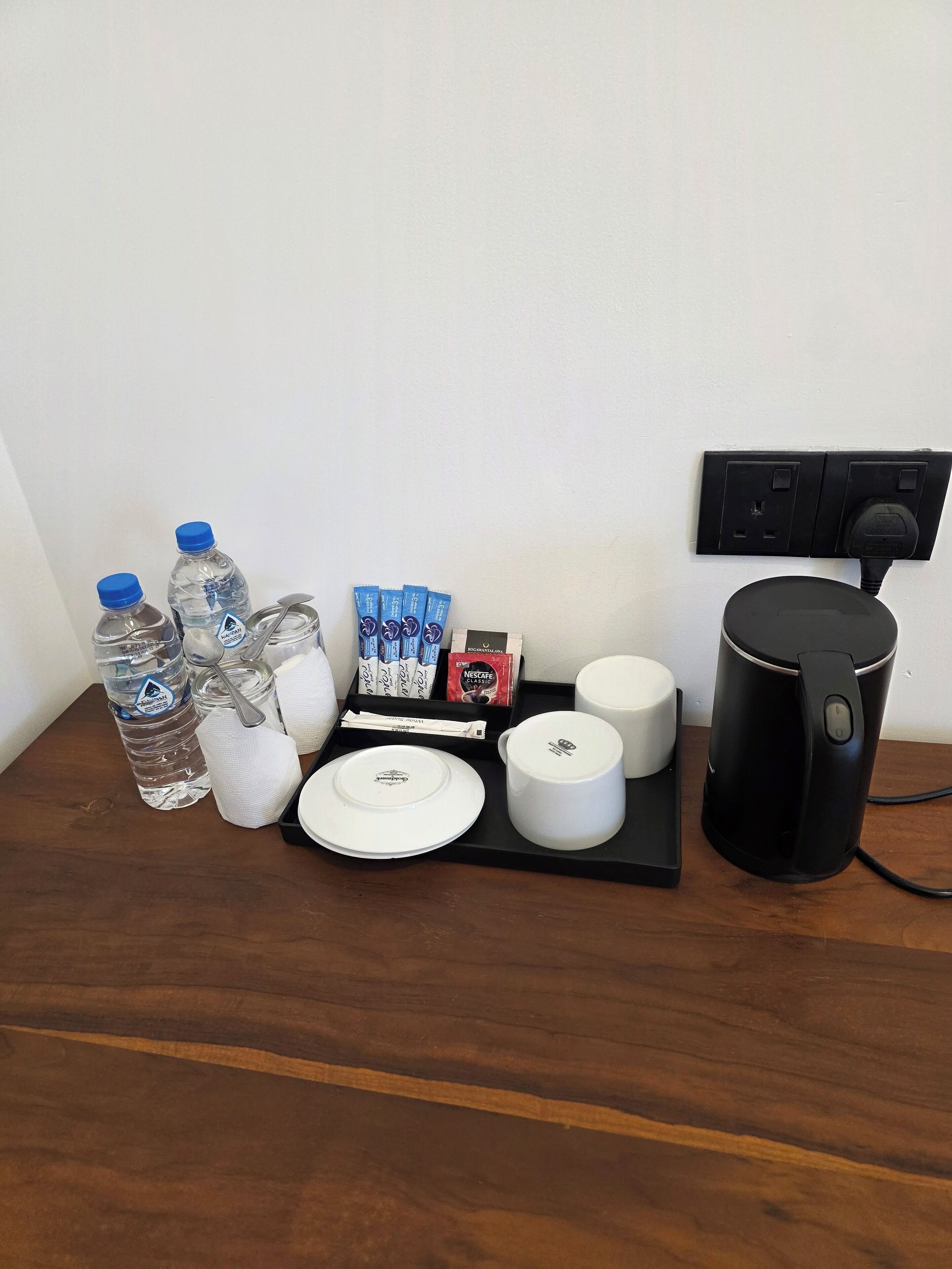 Une table sur laquelle se trouve un plateau noir, avec une bouteille d’eau, un verre, une cuillère, une assiette, une tasse, une machine à café et quelques sachets de café.
