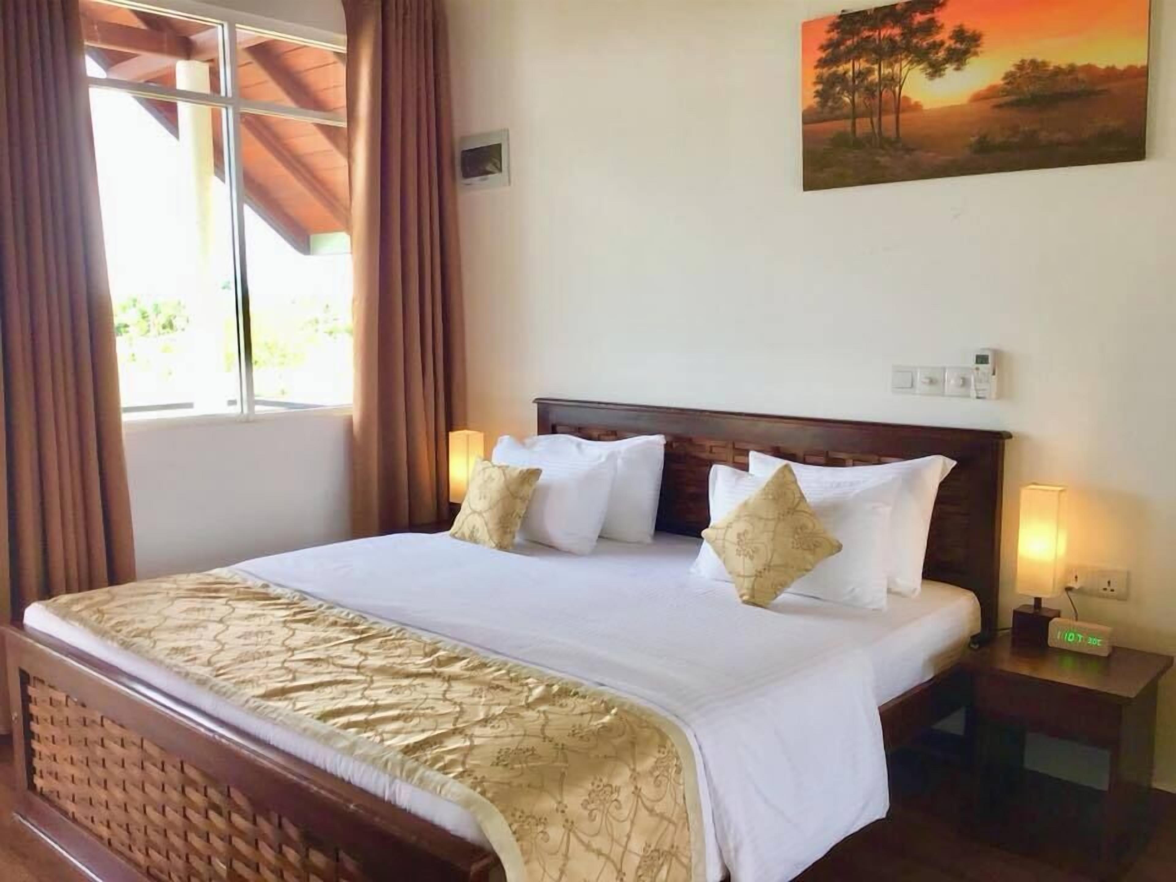 A neatly made bed with a patterned bedspread, two bedside lamps, and a painting of a sunset on the wall.