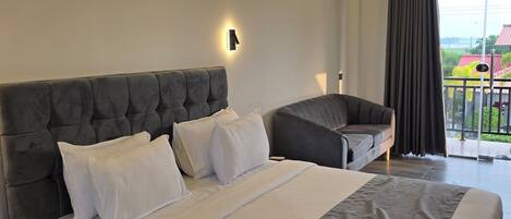 A modern hotel room with a large bed, a smaller sofa, a nightstand, and a view of the outdoors through a large window.