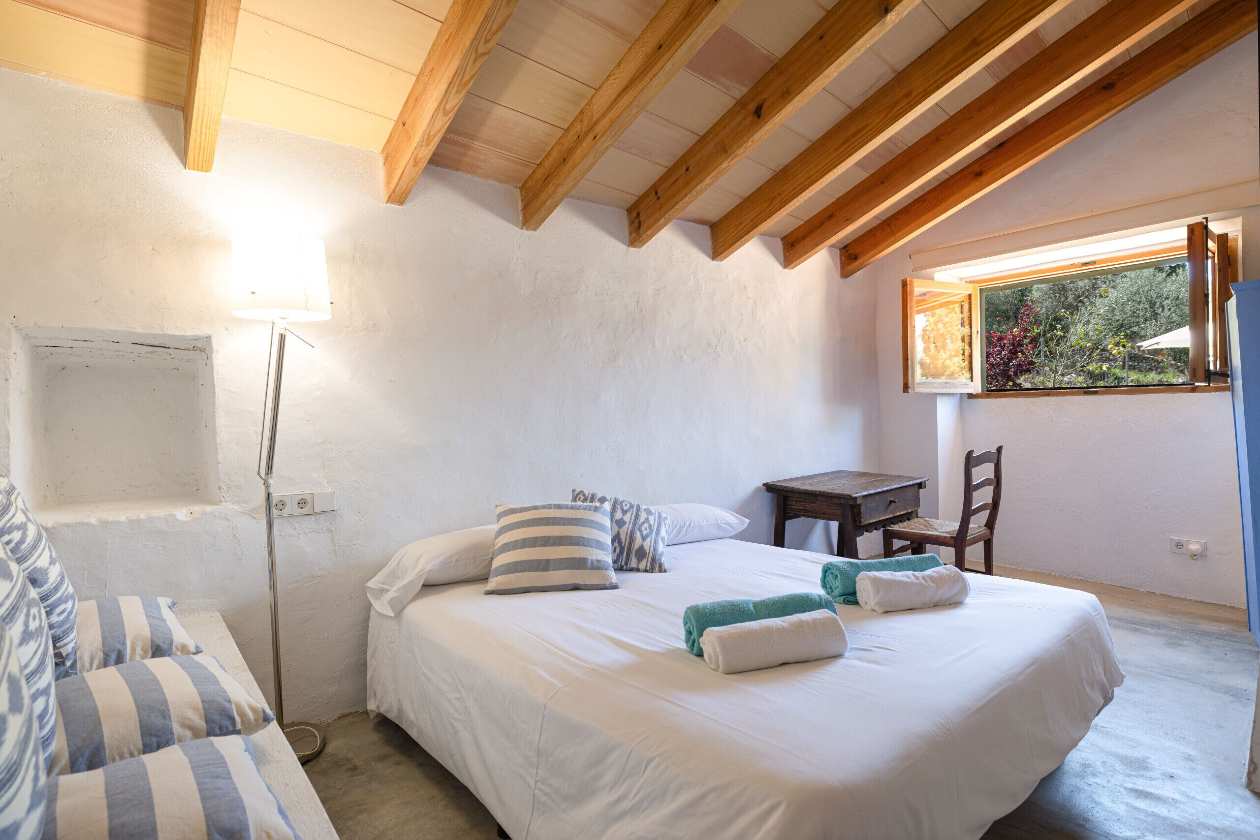 A bedroom with a bed, bedside table, chair, lamp, window, and wooden beams.