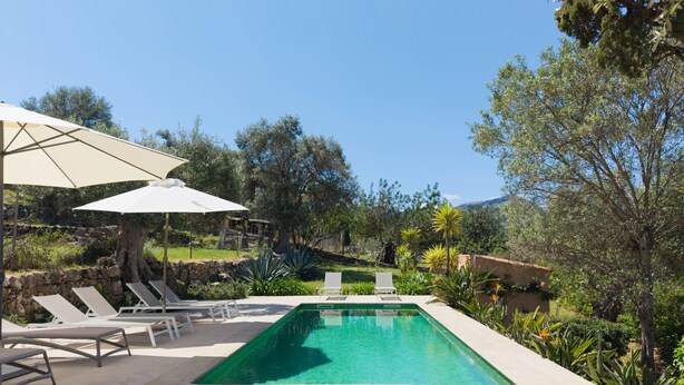A rectangular swimming pool with a clear blue water, surrounded by a stone wall, and equipped with lounge chairs and a large umbrella. - Finca Es Coste by Rentallorca (Pollensa)
