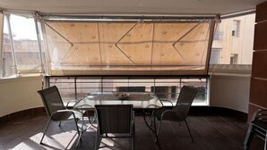 Ein Eckzimmer mit Tisch und vier Stühlen, ein großes Fenster mit Holzjalousie und Blick auf Gebäude.