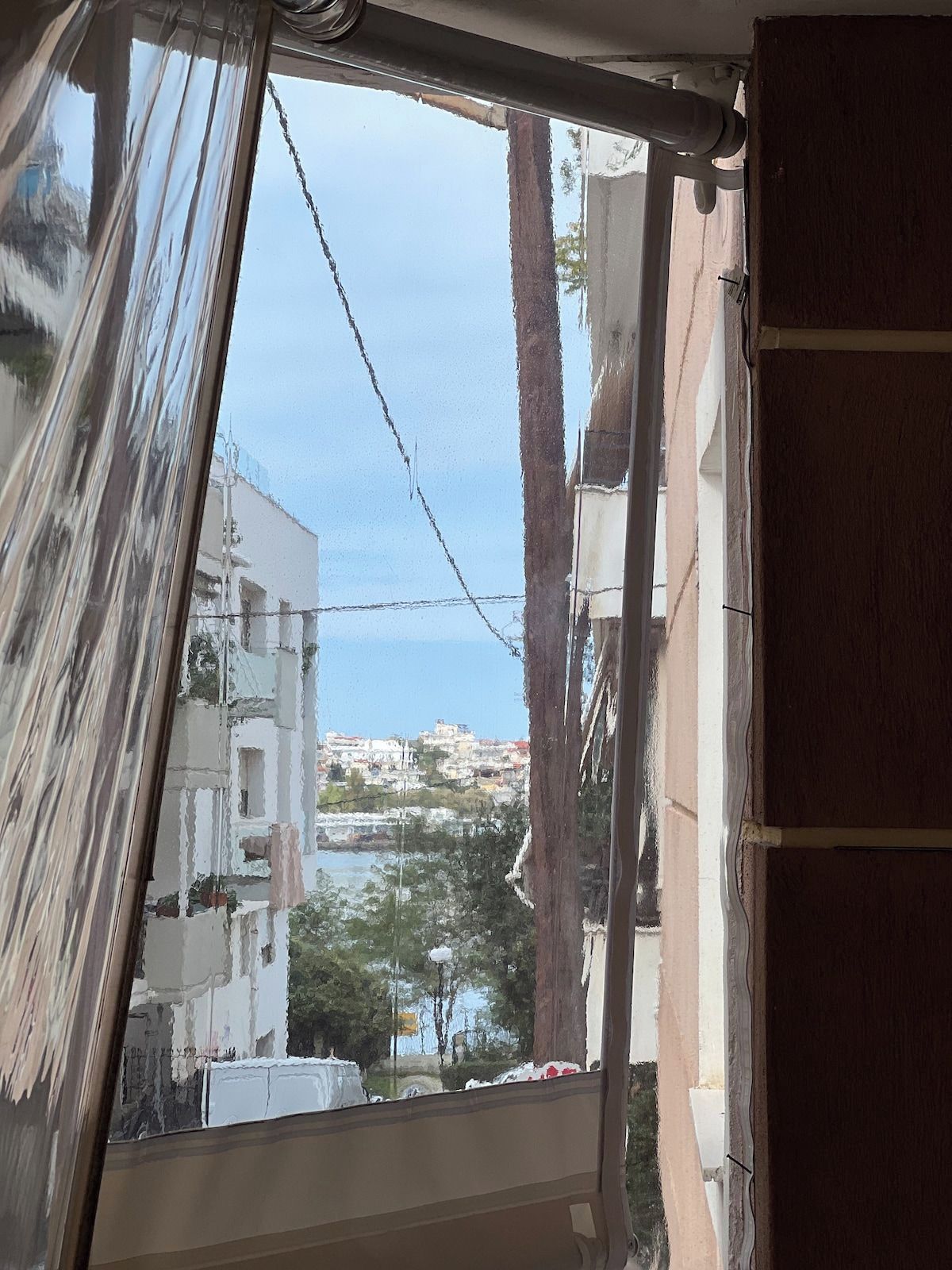 A window with a view of a coastal town, a tree, and a building with a balcony.