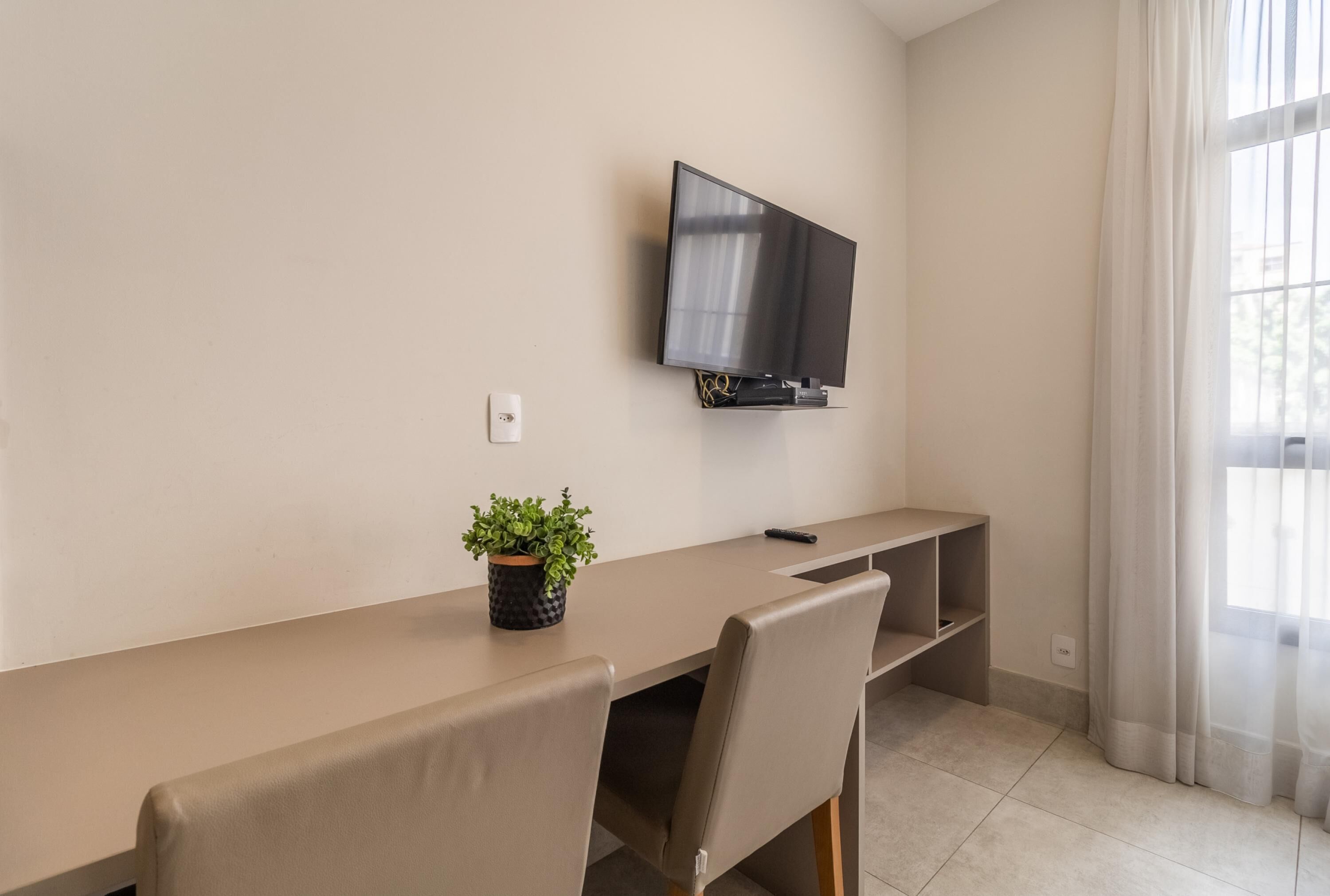 Una sala de estar moderna con un televisor de pantalla plana montado en la pared, una mesa larga con sillas, una pequeña planta en maceta y una ventana con cortinas traslúcidas.