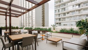 An outdoor patio area with tables and chairs, surrounded by greenery and overlooking a residential building.