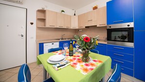 A compact kitchen with a dining area, featuring a table set for two, a wine bottle, and a vase with flowers. - Casa Blu (Toscolano Maderno)
