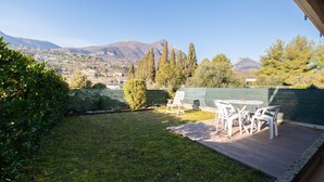 A patio with a table and chairs, a small lawn, and a view of a town and mountains in the distance. - Casa Blu (Toscolano Maderno)
