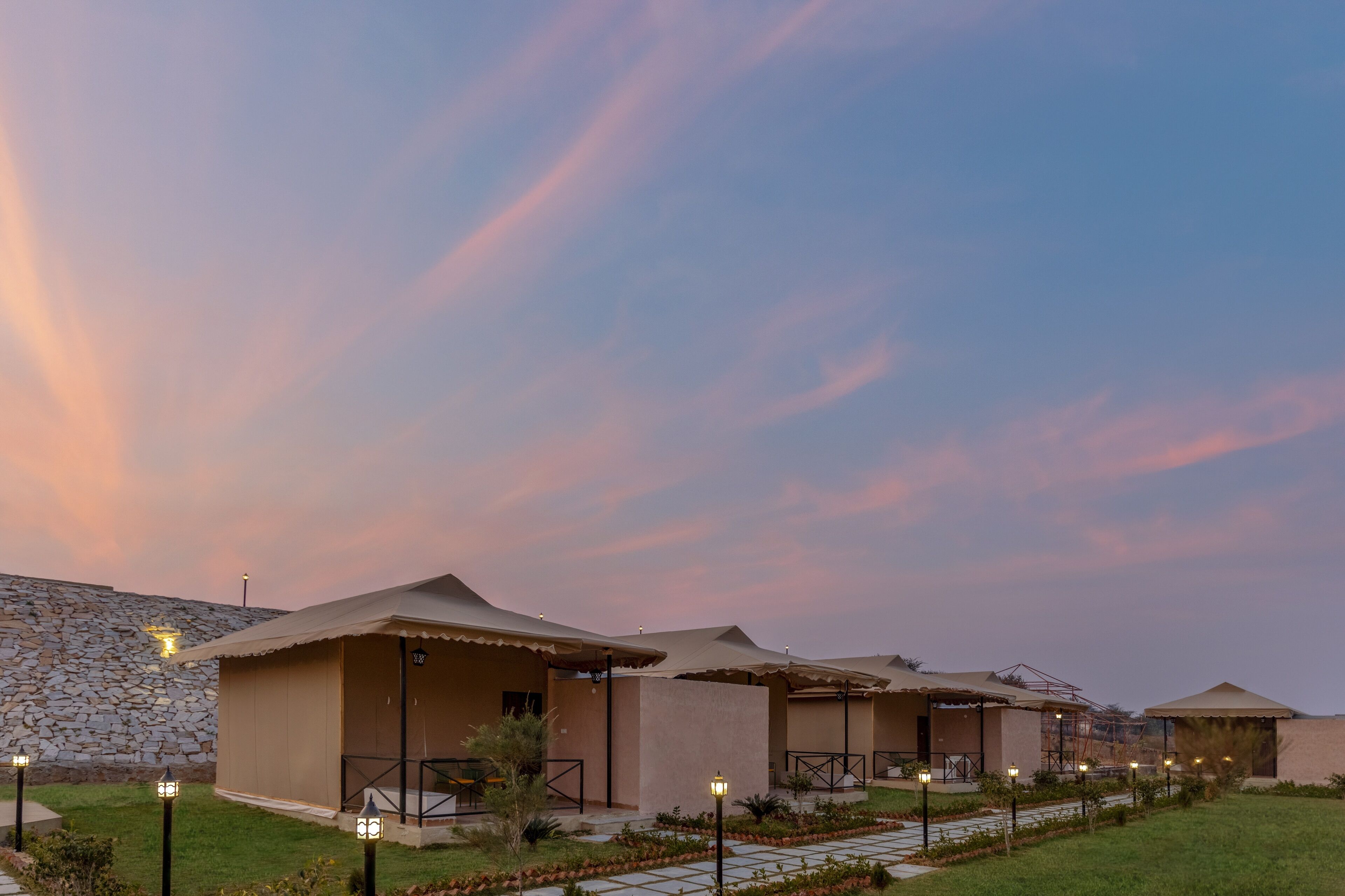 A row of modern, single-story buildings with flat roofs, arranged in a linear fashion along a stone wall. Each building has a covered outdoor area with a table and chairs.