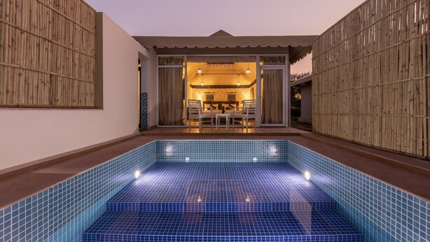 A swimming pool with blue tiles, a dining area with a table and chairs, and a covered seating area with a view of the pool. - Sterling ReWild Sariska (Thanagazi)
