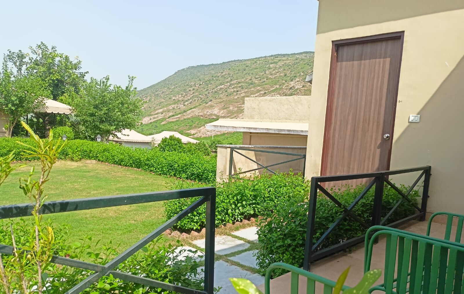 A balcony with a metal railing, a door, and a view of a landscaped garden with bushes and a hill in the background.