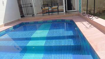 Una piscina rettangolare con piastrelle blu, una zona prendisole con tavolo e sedie, e una recinzione in metallo.