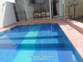 Una piscina rectangular con azulejos azules, un patio con mesa y sillas, y una cerca de metal.