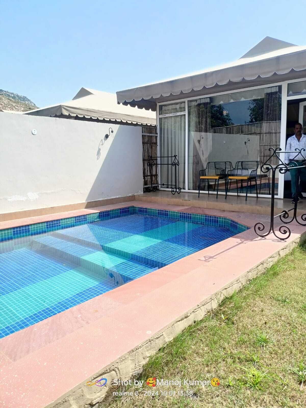 A swimming pool with a tiled deck, a covered patio area with outdoor seating, and a white exterior wall.