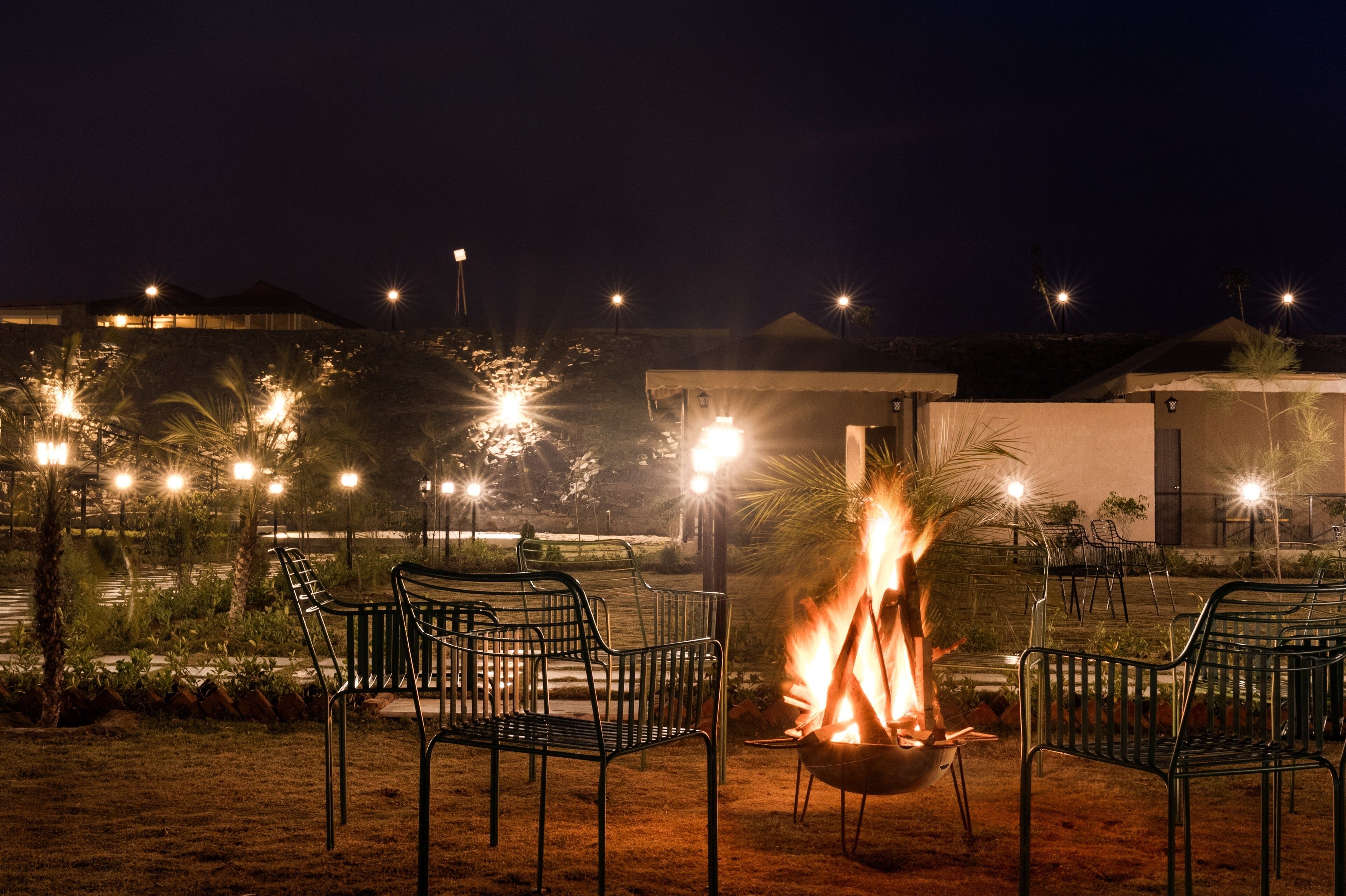 A nighttime outdoor seating area with a fire pit, surrounded by metal chairs and illuminated by ambient lighting.