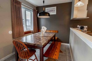 Un espace repas comprenant une table, des chaises, un banc, un luminaire suspendu, une fenêtre avec des rideaux et un comptoir de cuisine équipé d’appareils électroménagers.