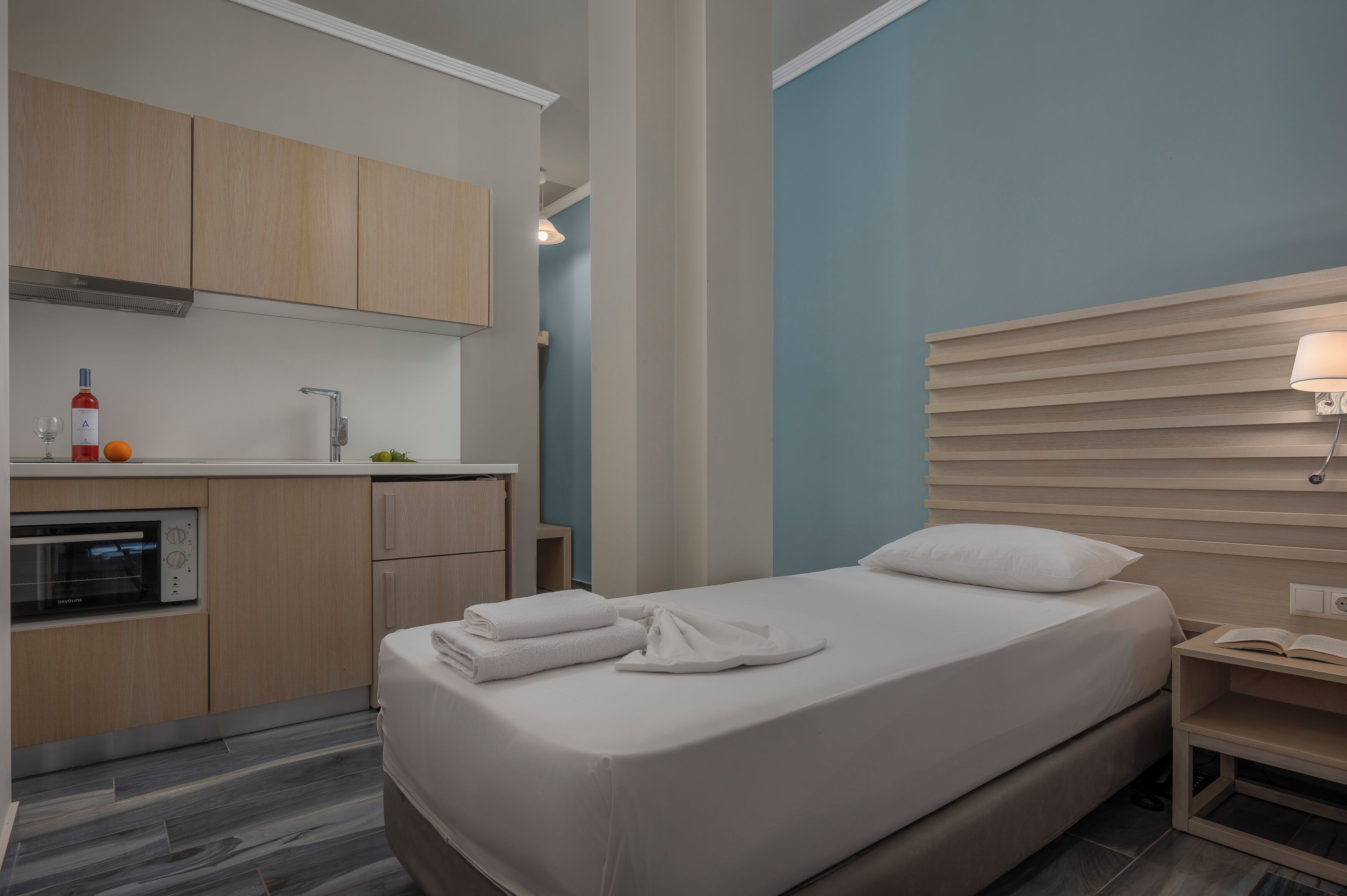 A modern hotel room with a bed, a bedside table, a nightstand, a lamp, a small table with a book, a kitchen area with a microwave, and a sink.