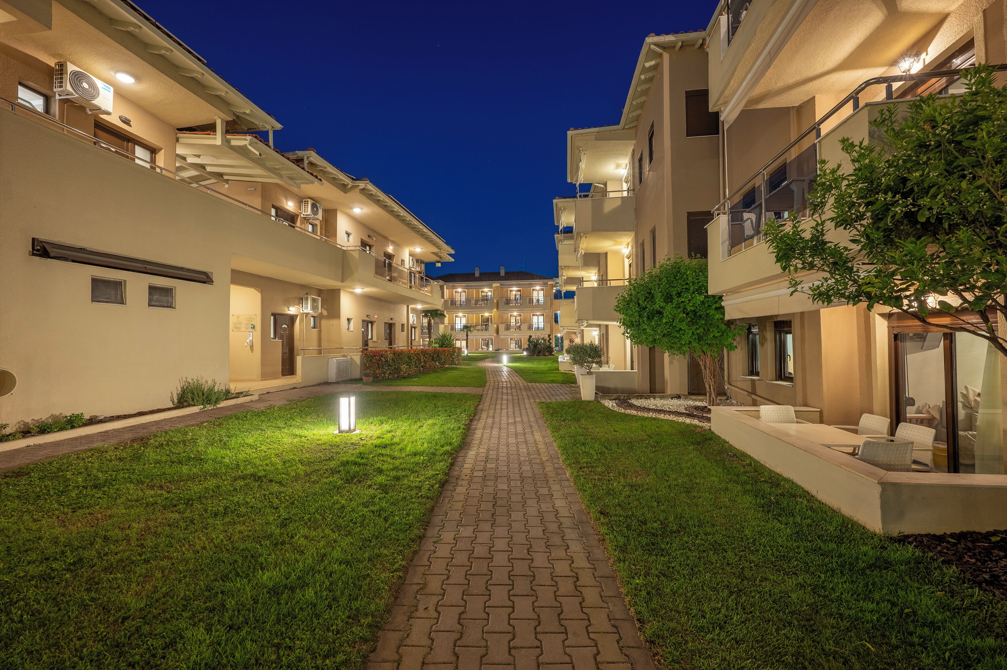 A well-lit courtyard with a paved walkway, surrounded by multi-story buildings with balconies and outdoor seating areas.