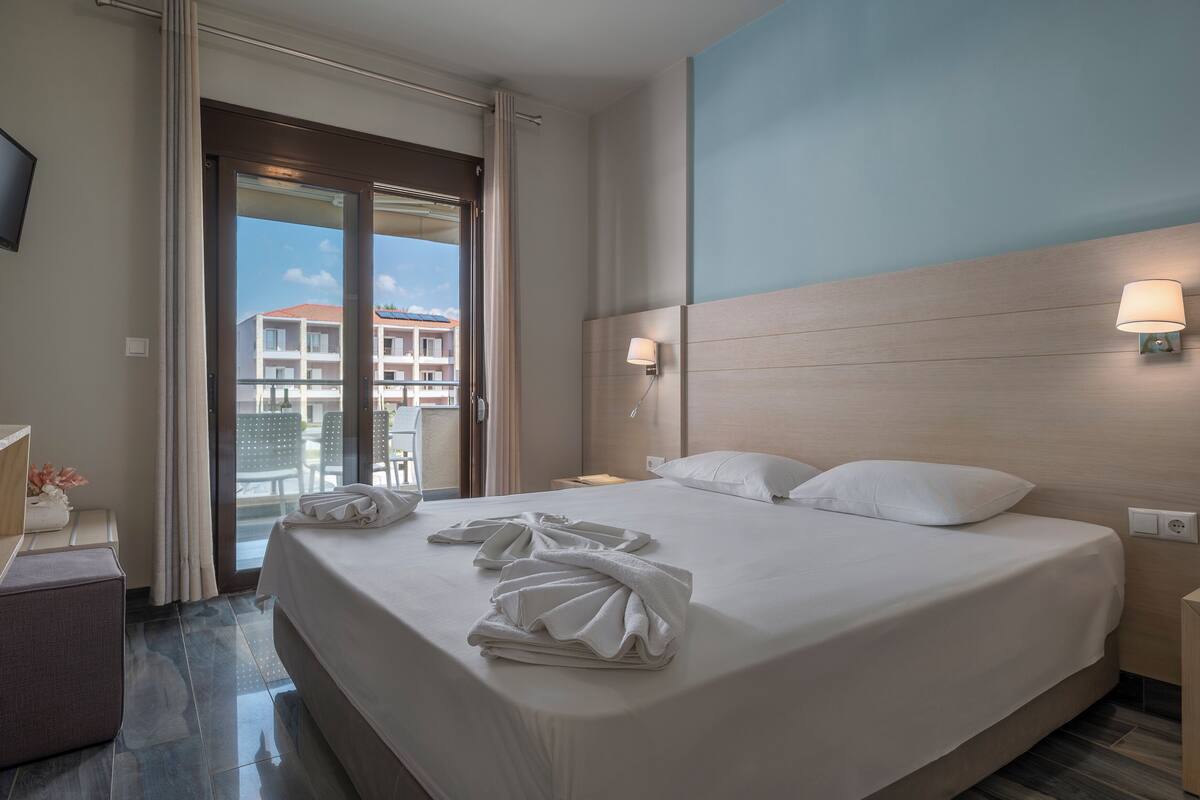 Ein Hotelzimmer mit einem großen Bett, zwei Nachttischlampen, einem Nachttisch, einem Schreibtisch mit Lampe und Blick auf den Balkon.