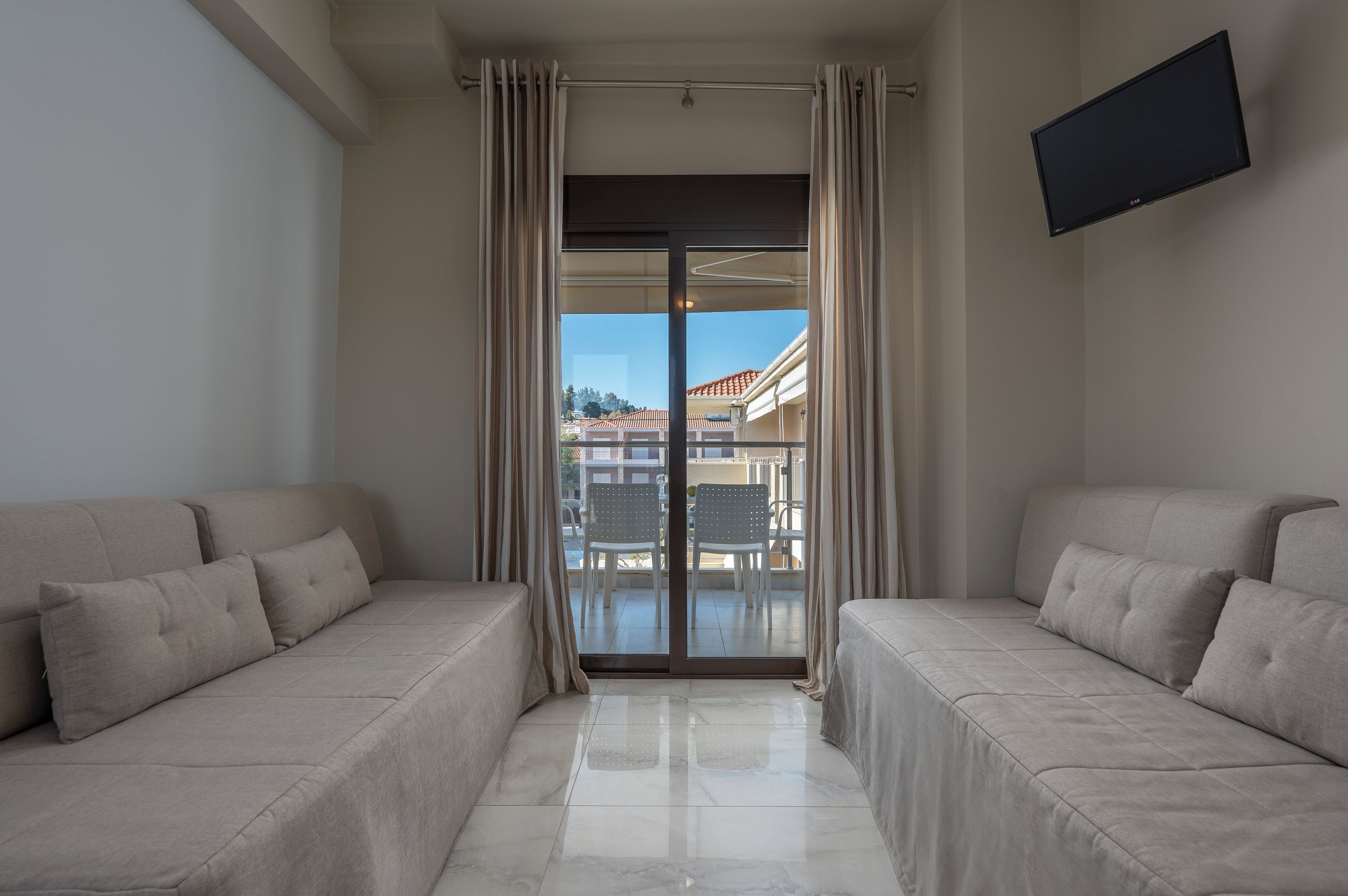 A modern hotel room with two beige sofas, a flat-screen TV, and a balcony with a view of buildings and a clear sky.