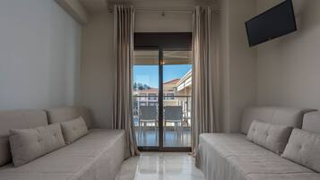 A modern hotel room with two beige sofas, a flat-screen TV, and a balcony with a view of buildings and a clear sky.