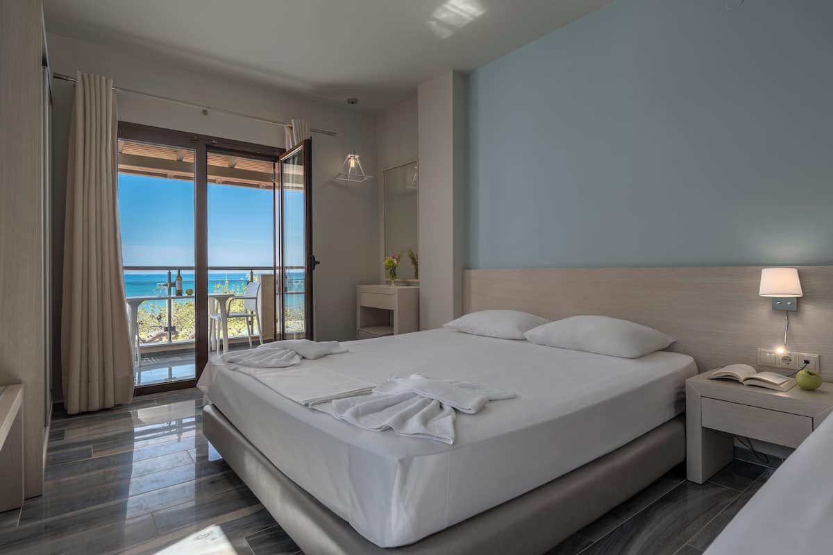 Ein modernes Hotelzimmer mit einem großen Bett, einem Nachttisch mit Lampe, Meerblick und einem Balkon mit Tisch und Stühlen.