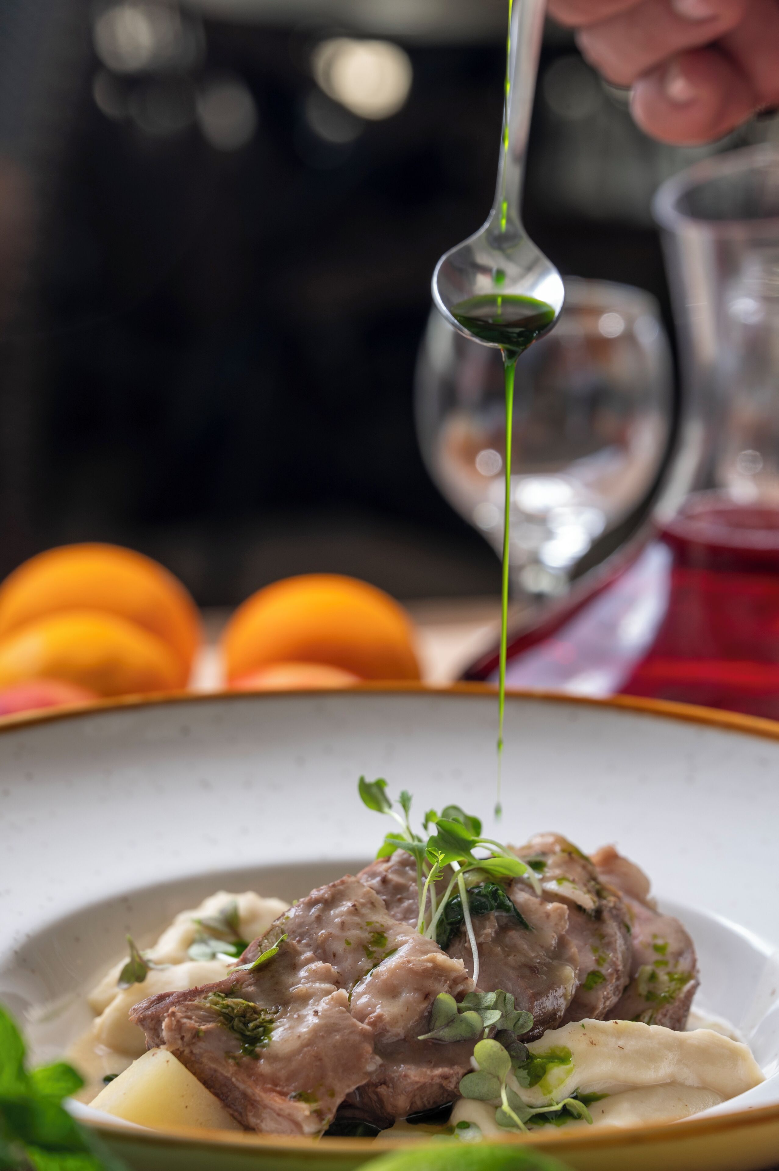 A close-up of a dish with meat and vegetables, garnished with greens, and a spoon pouring a green sauce over it.