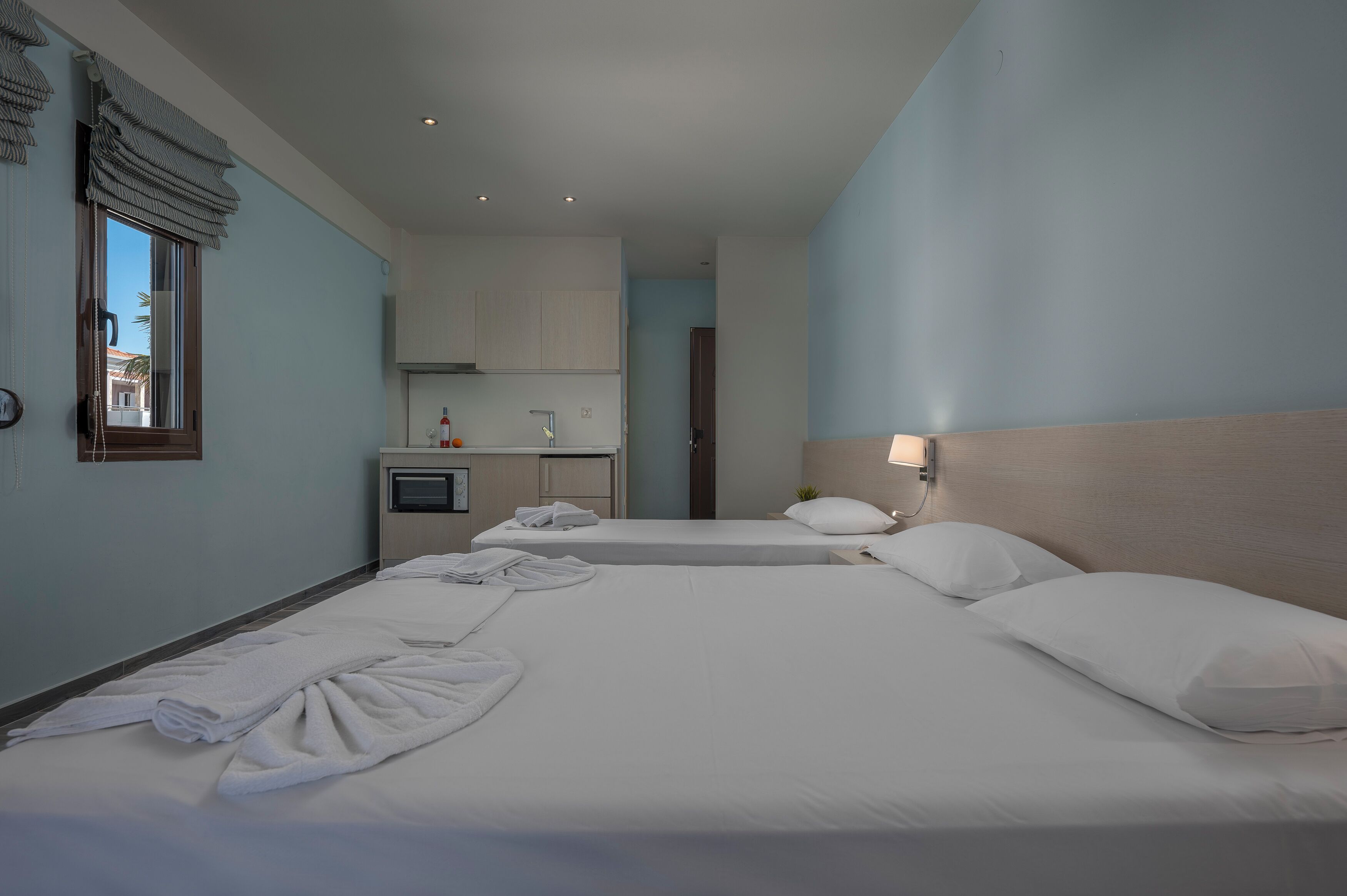 A hotel room with two beds, a small kitchenette, and a window with curtains.